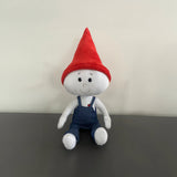 The Gnome Doll is a white stuffed doll with a red pointy gnome hat and blue denim overalls. It is sitting on a gray counter top in front of a white wall.