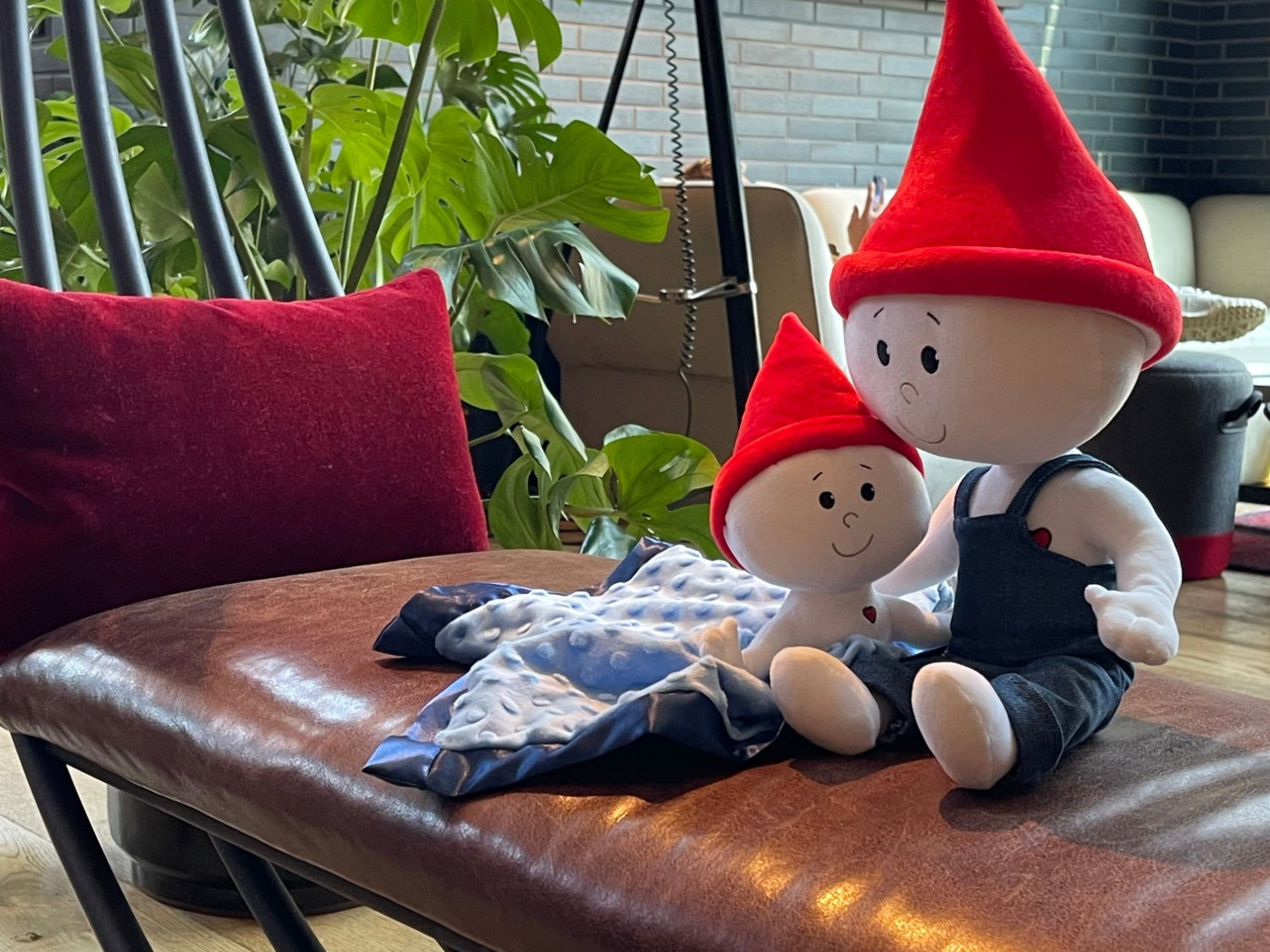 The Gnome Doll and Gnome Lovey are sitting together on a brown leather bench in a family's living room. The gnome doll is a soft stuffed doll that is white with a red pointy gnome hat and blue denim overalls. The gnome lovey is a soft stuffed doll that is white but is attached to a light blue blanket with navy blue trim. The lovey also has a red pointy gnome hat.