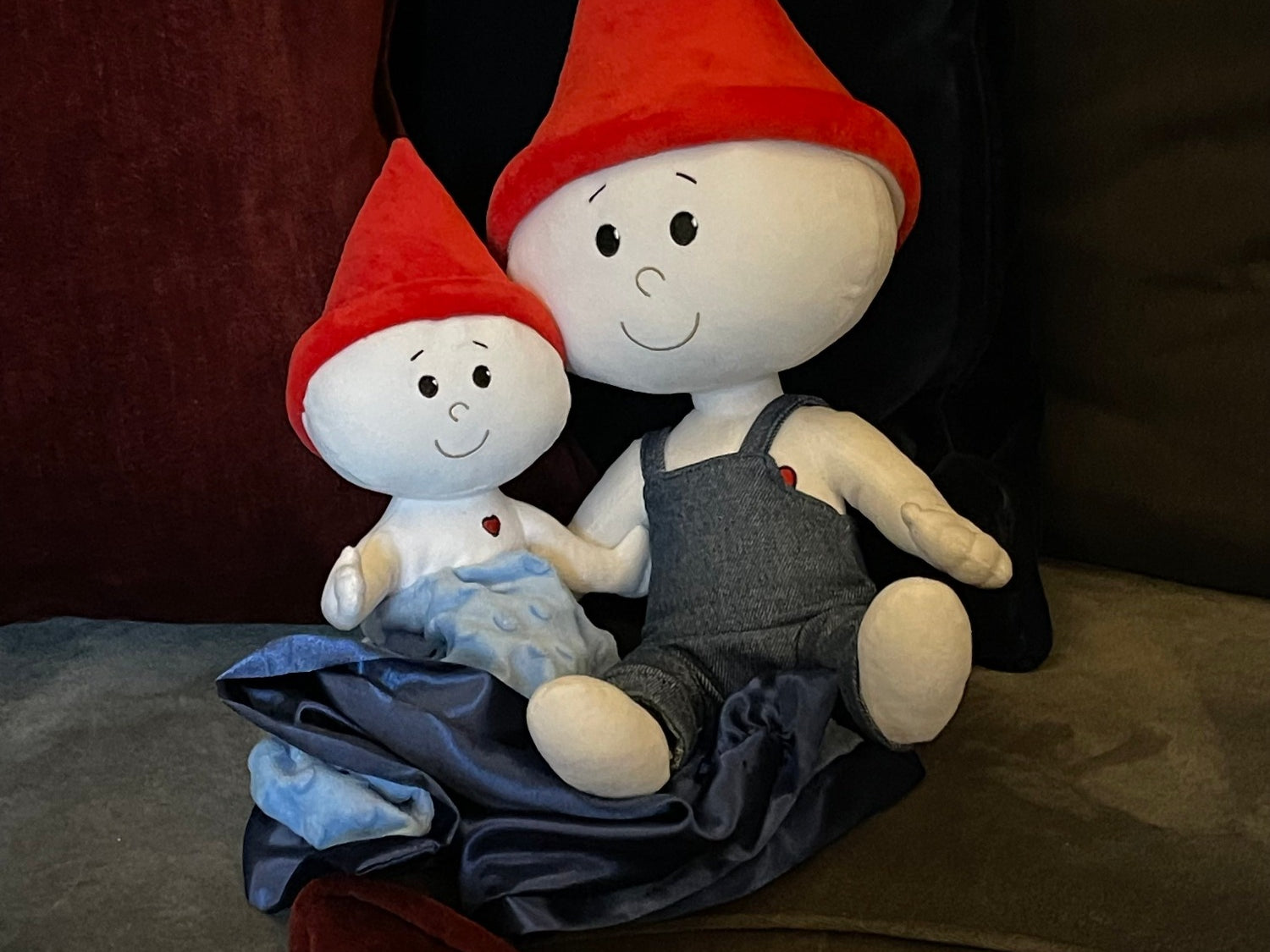 The Gnome Doll and Gnome Lovey are sitting together on a gray couch in a family's living room. The gnome doll is a soft stuffed doll that is white with a red pointy gnome hat and blue denim overalls. The gnome lovey is a soft stuffed doll that is white but is attached to a light blue blanket with navy blue trim. The lovey also has a red pointy gnome hat.