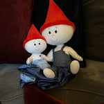 The Gnome Doll and Gnome Lovey are sitting together on a gray couch in a family's living room. The gnome doll is a soft stuffed doll that is white with a red pointy gnome hat and blue denim overalls. The gnome lovey is a soft stuffed doll that is white but is attached to a light blue blanket with navy blue trim. The lovey also has a red pointy gnome hat.