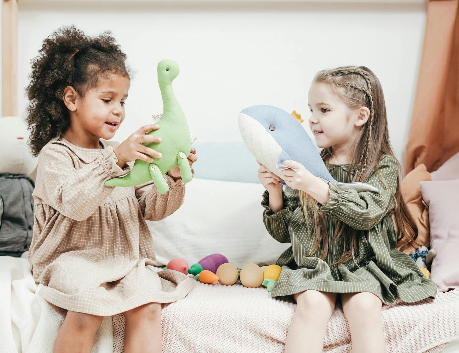 Two young children sit cross-legged on a cozy, textured throw, joyfully playing with plush toys—a green dinosaur and a blue whale—against a background of soft pastels and playful decor.