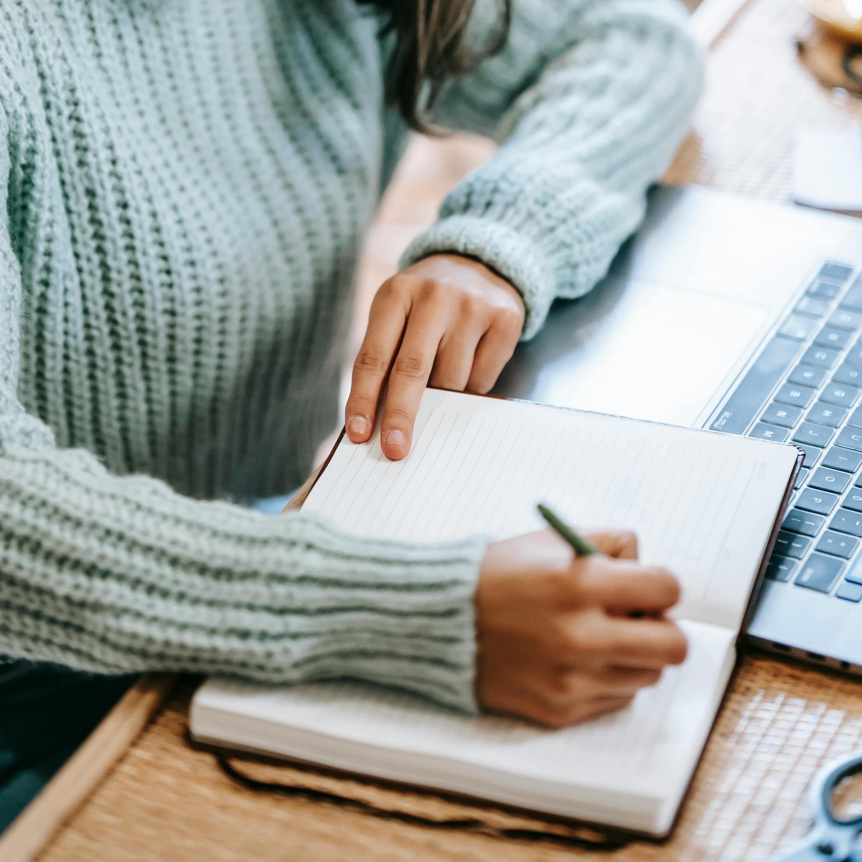 A close-up of a person's hand holding a pen, poised to write in a notebook beside a laptop, indicating a moment of thought or planning.