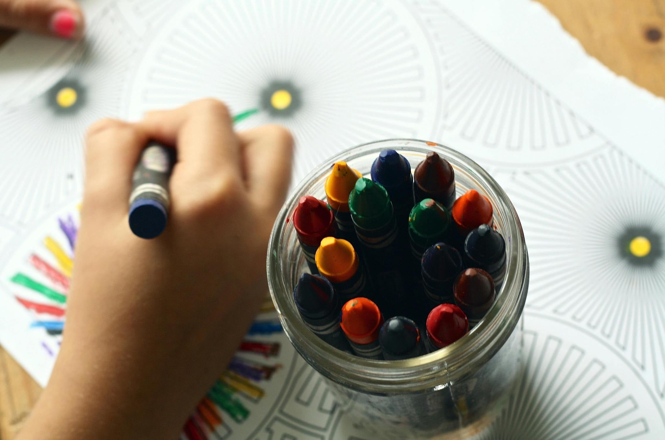 Overhead shot of a child's hand holding a blue crayon, coloring in a detailed mandala design, with a jar full of vibrant crayons in the foreground, on a wooden surface.