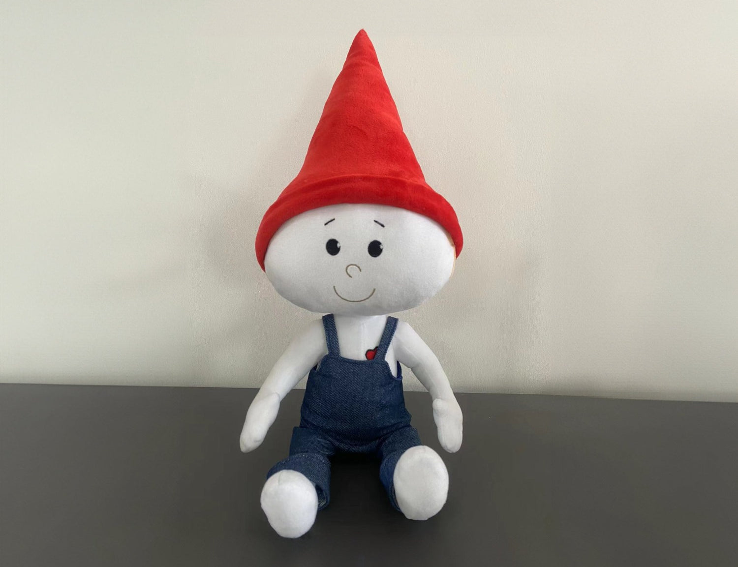 The Gnome Doll is a white stuffed doll with a red pointy gnome hat and blue denim overalls. It is sitting on a gray counter top in front of a white wall.