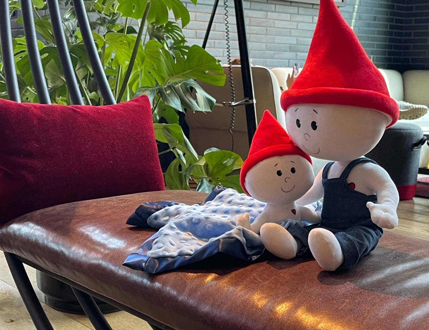 The Gnome Doll and Gnome Lovey sitting together on a brown leather bench in a family's living room. The doll is white with a red pointy gnome hat and denim overalls. The gnome lovey is a smaller white stuffed doll with a red pointy hat and is attached to a light blue blanket with navy blue trim.