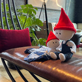 The Gnome Doll and Gnome Lovey sitting together on a brown leather bench in a family's living room. The doll is white with a red pointy gnome hat and denim overalls. The gnome lovey is a smaller white stuffed doll with a red pointy hat and is attached to a light blue blanket with navy blue trim.