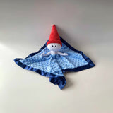 The Gnome Lovey is a stuffed doll attached to a light blue blanket with navy blue trim. The lovey's face and body is white, with a tall red gnome hat and a small red heart embroidered on the left side of it's chest.