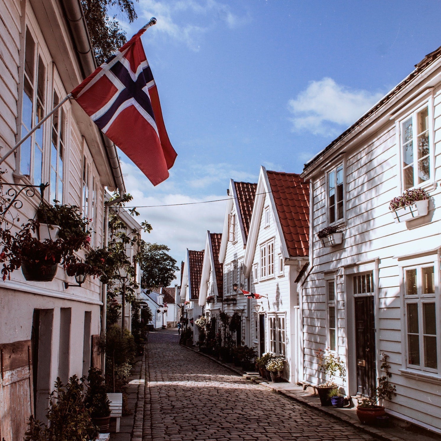 A quaint cobblestone street lined with traditional white wooden houses under a clear blue sky. A vibrant Norwegian flag waves proudly from one of the houses, adding a splash of red, blue, and white to the charming scene.