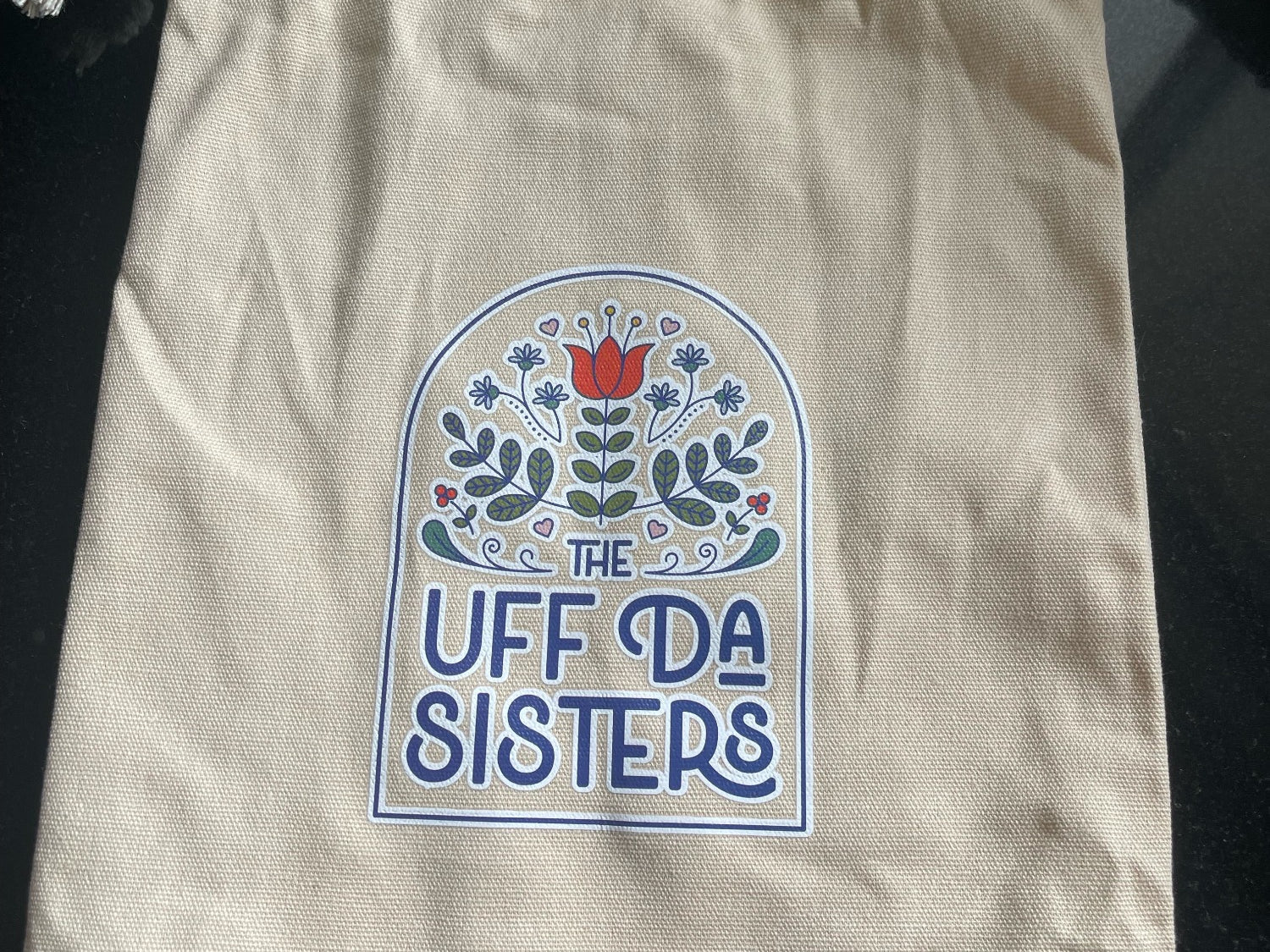 A natural-colored canvas drawstring bag with the logo of The Uff Da Sisters printed on the front. The logo features a stylized floral design in a circular motif with the text 'THE UFF DA SISTERS' around it. The bag appears to be designed for holding items like the brand's Gnome Doll or Gnome Lovey.