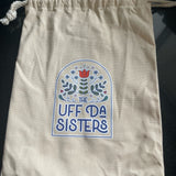 A natural-colored canvas drawstring bag with the logo of The Uff Da Sisters printed on the front. The logo features a stylized floral design in a circular motif with the text 'THE UFF DA SISTERS' around it. The bag appears to be designed for holding items like the brand's Gnome Doll or Gnome Lovey.