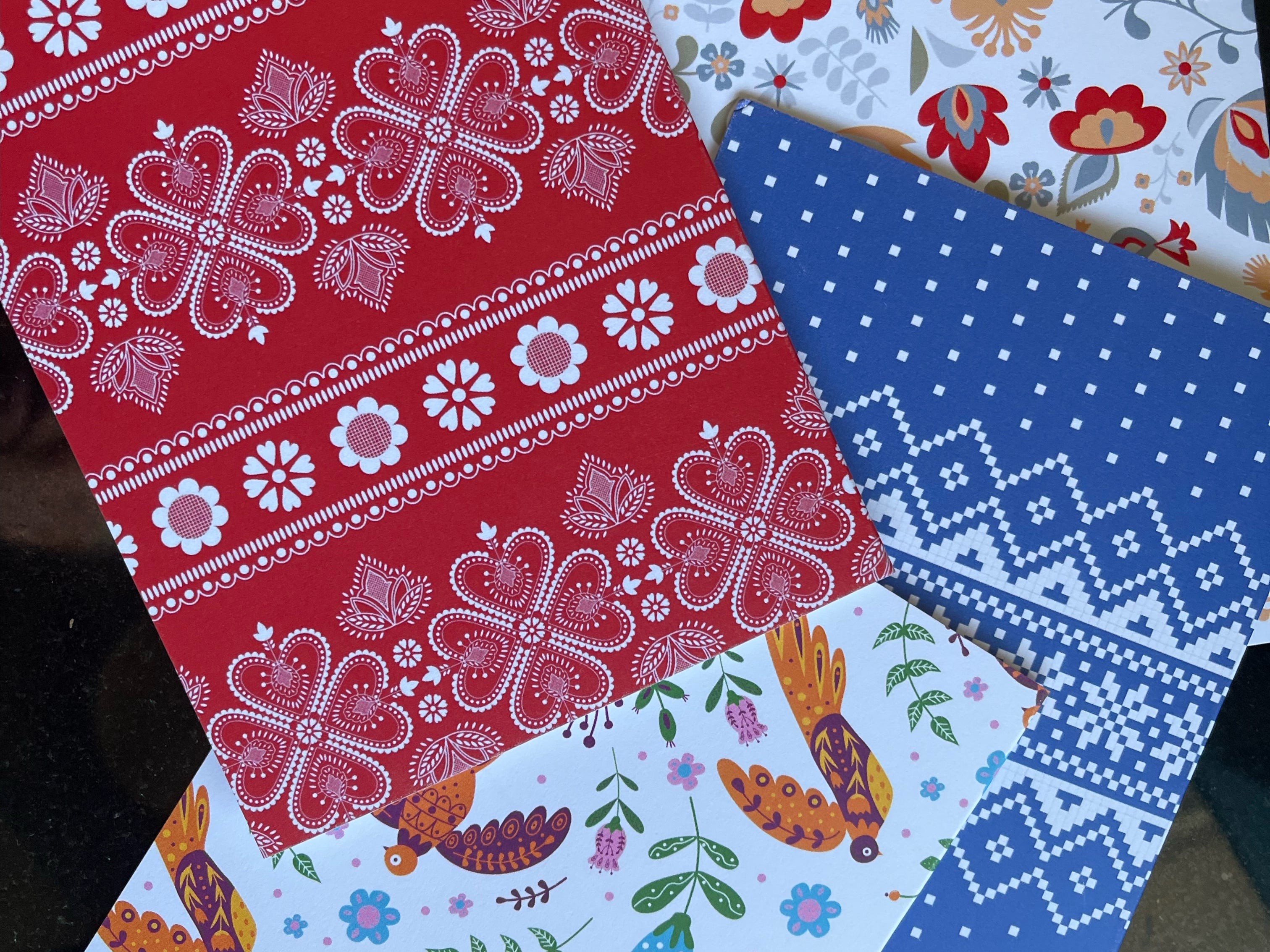 A collection of four notecards with Nordic designs from The Uff Da Sisters, featuring patterns in red with white floral motifs, grey with multicolored flowers, white with colorful birds and floral designs, and blue with white geometric patterns.