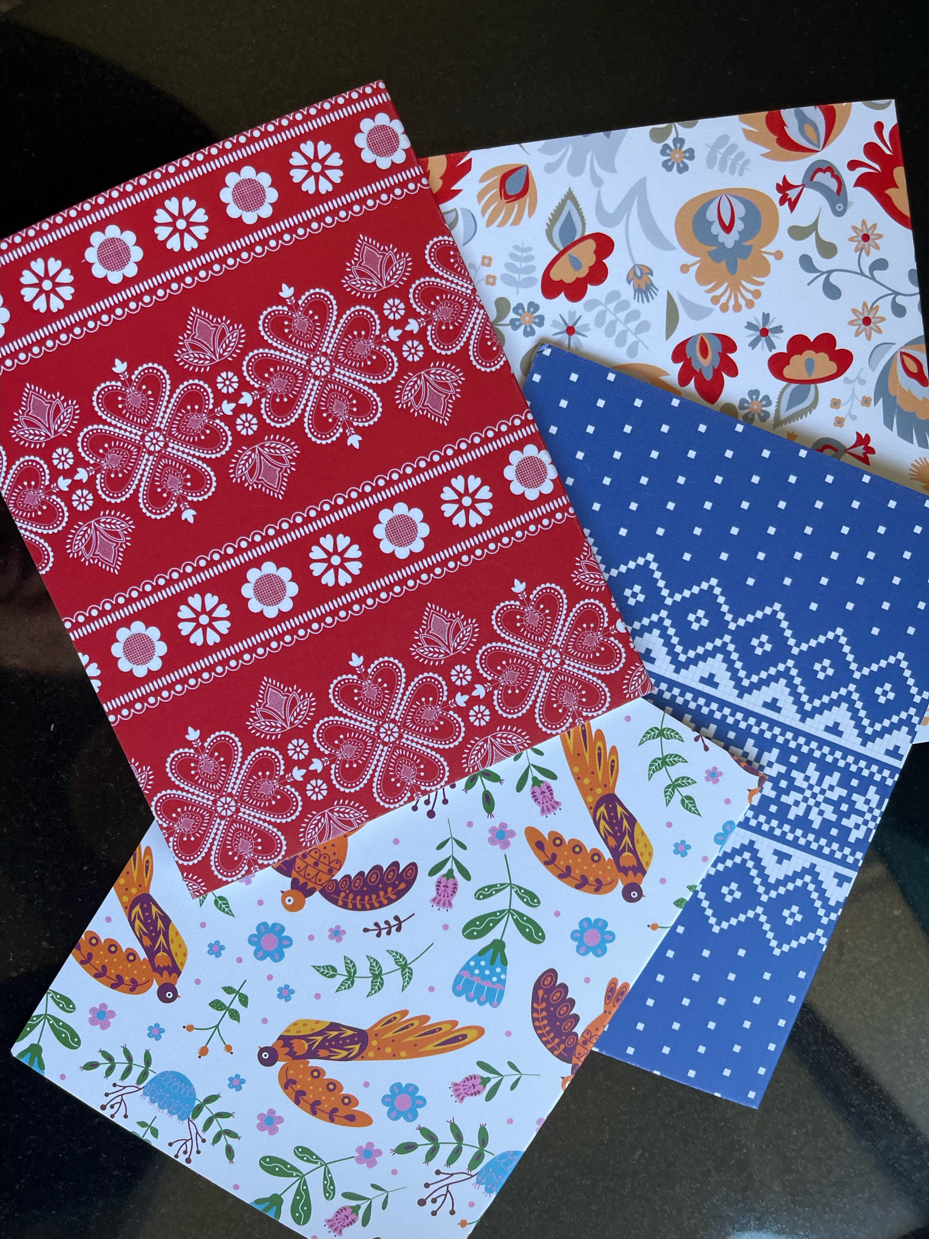 A collection of four notecards with Nordic designs from The Uff Da Sisters, featuring patterns in red with white floral motifs, grey with multicolored flowers, white with colorful birds and floral designs, and blue with white geometric patterns.